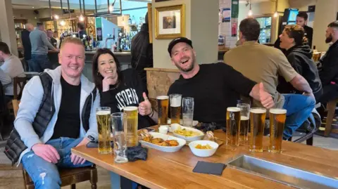 Gedling Inn Три человека сидели за столом в пабе, улыбаясь и с большими пальцами, с многочисленными пивами перед ними