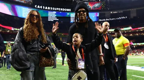 Reuters Blue Ivy носит негабаритную кожаную куртку в паре с повседневной грузовой джинсовой ткани; стоя рядом с отцом Джеем З. и сестрой Руми