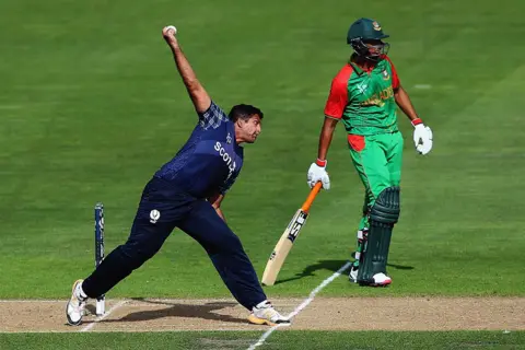 Getty Images Majid Haq из Scotland Bowls во время матча чемпионата мира по крикету ICC 2015 года между Бангладеш и Шотландией на Saxton Field 5 марта 2015 года в Нельсоне, Новая Зеландия. Он на бок, надев синий и боулинг полного шкура в другой калипе слева направо от выстрела. Игрок Бенгладеши в зеленом и красном стоит позади него на другой калитки.