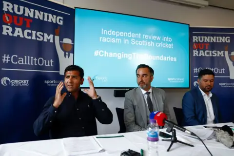 Getty Images Majid Haq и Kasim Sheik проводят пресс -конференцию с адвокатом Аамера Анвара в отеле Stirling Court 25 июля 2022 года в Стерлинге. Они одеты небрежно в темно-синие и белые рубашки с открытыми шеей, а мистер Анвар находится посередине в сером костюме с тремя частями. На столе есть микрофон, заканчивая за ними фирменные доски расизма.