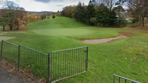 Google Общее представление о футболке на поле для гольфа Dalmuir. На переднем плане изображения есть серый забор. На тройнике гольф в темной одежде размахивает клубом. Он окружен зеленью на курсе.
