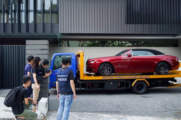 Красная Rolls-Royce загружается на фальшивый эвакуатор перед современным зданием, с несколькими людьми, включая полицейского, наблюдение и документирование сцены