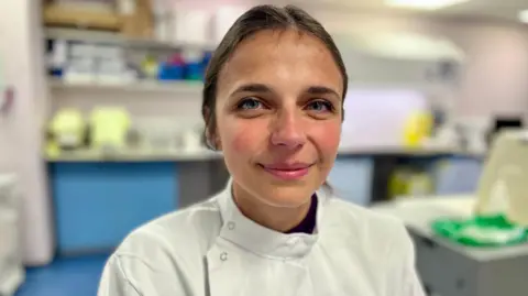 Никки Фокс/BBC Dr Maura Malpetti из Кембриджского университета смотрит на камеру. Она стоит в лаборатории и имеет белый лабораторный халат. Вы можете видеть ее голову и плечи, и она улыбается. Вы можете разобрать некоторые полки, размытые на фоне.