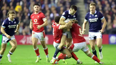 PA Media Scotland Player Blair Kinghorn, одетый в Шотландскую полосу голубых майки и белых шорт, решается двумя игроками Уэльса в красных майках и белых шортах, во время матча Six Nations. Игроки обеих команд смотрят на заднем плане.  