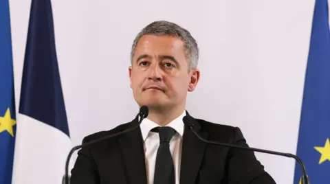 EPA Gérald Dermanin (мужчина среднего возраста с седыми волосами с короткими сжиманием) стоит перед двумя микрофонами на подиуме в черном костюме, белой рубашке и черном галстуке. За ним находятся два флага ЕС и французский с нижней частью рамки