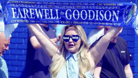 PA Media, фанат Everton с длинными светлыми волосами за землей, держащий шарф в воздухе, в котором говорится «Прощай Гудсон»