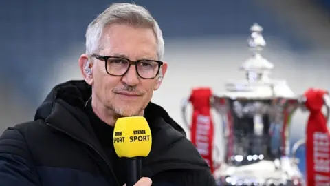 Getty Images Gary Lineker в толстом пальто, удерживающем желтый спортивный микрофон BBC, с чашкой анимации рядом с ним в 2021 году