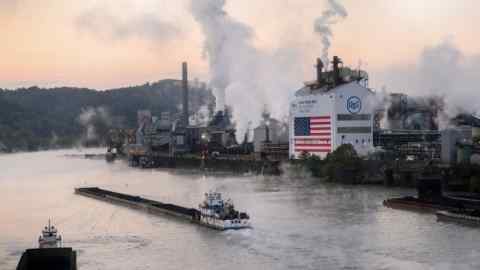 Буксир толкает баржу вдоль реки возле завода US Steel Corp в Клэртоне. Промышленная площадка излучает дым, а на здании выставлена ​​американский флаг.