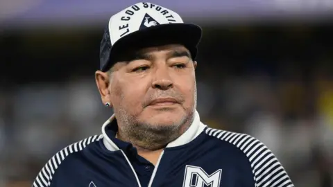 Getty Images Diego Maradona носит военно -морской и белую кепку и соответствующую куртку на футбольном матче в Буэнос -Айресе. У него есть правое ухо, и он отводит взгляд от камеры.
