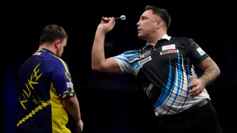 PA Media Gerwyn Price, на фото бросал дротик в дротику с выстрела. На нем черная рубашка с синими и белыми осколками и белым воротником. У Люка Литтлера его спина к камере, смотрит прямо в будущее. Фон черный.