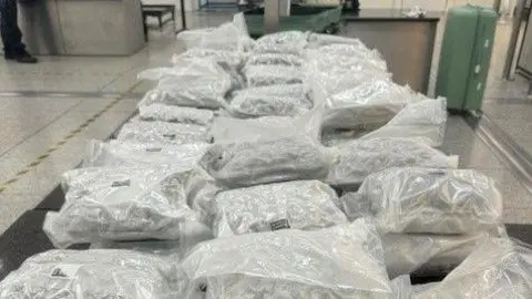 NCA Много вакуумных запечатанных белых пластиковых пакетов, выложенных на столе. Два зеленых чемодаза находятся на заднем плане в зале аэропорта.