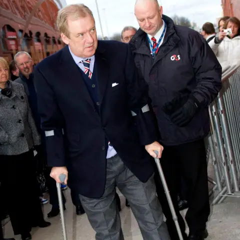 SNS, сэр Дэвид Мюррей на улице Ibrox в 2011 году. Он носит темный пиджак, светло -голубую рубашку и красный и синий полосатый галстук. Он ходит на костылях. Охранник стоит за ним.